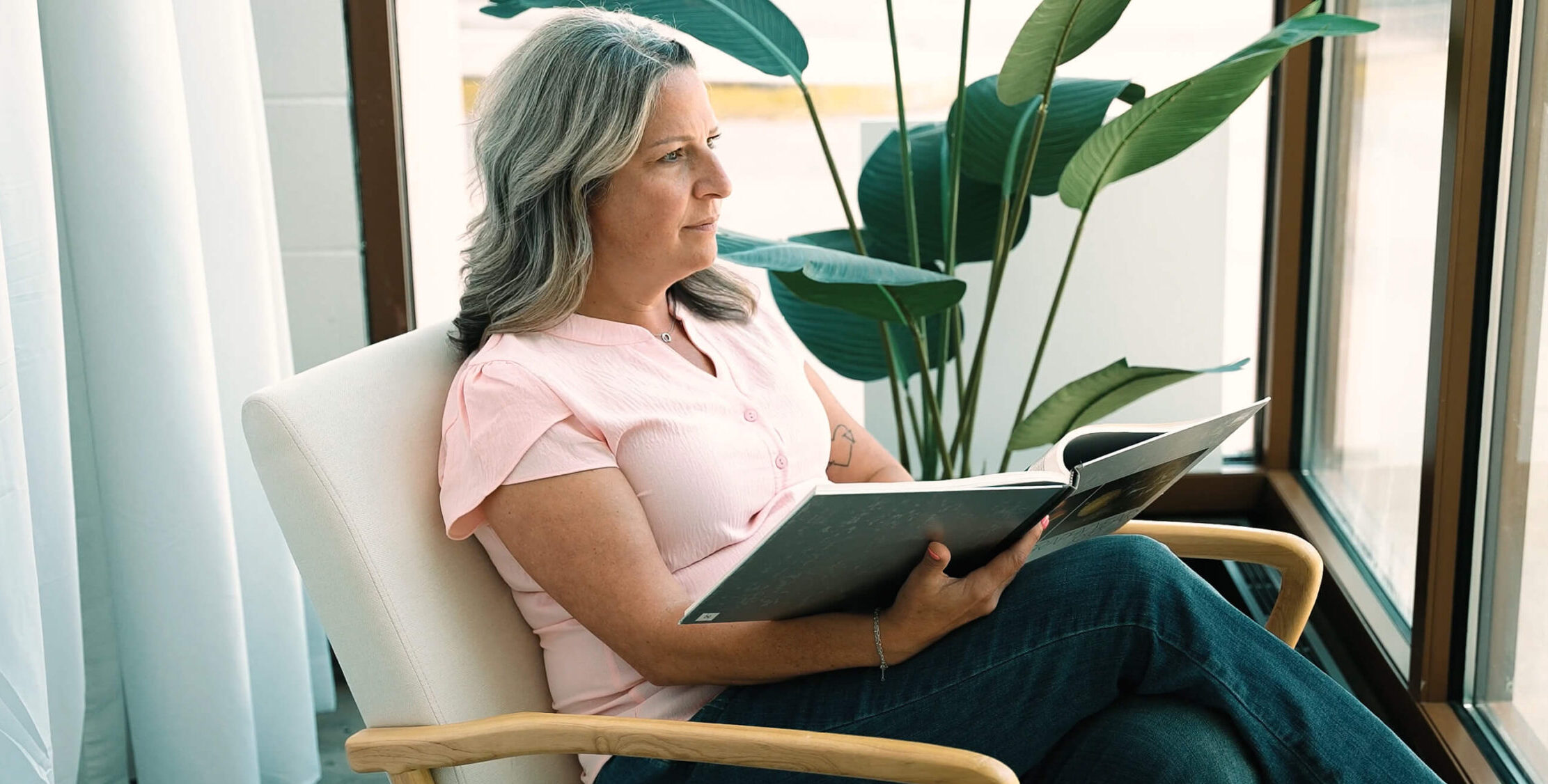 Kristin Miller, a breast cancer survivor and mammography tech at Envision Radiology sits by a window with a book in her lap.